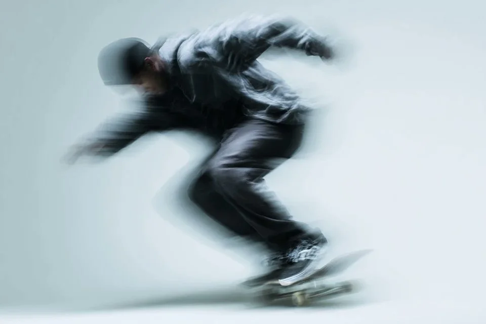 A minimalist sport advertising image of a skateboarder in dark clothing captured mid-motion with heavy blur against a pale neutral background.