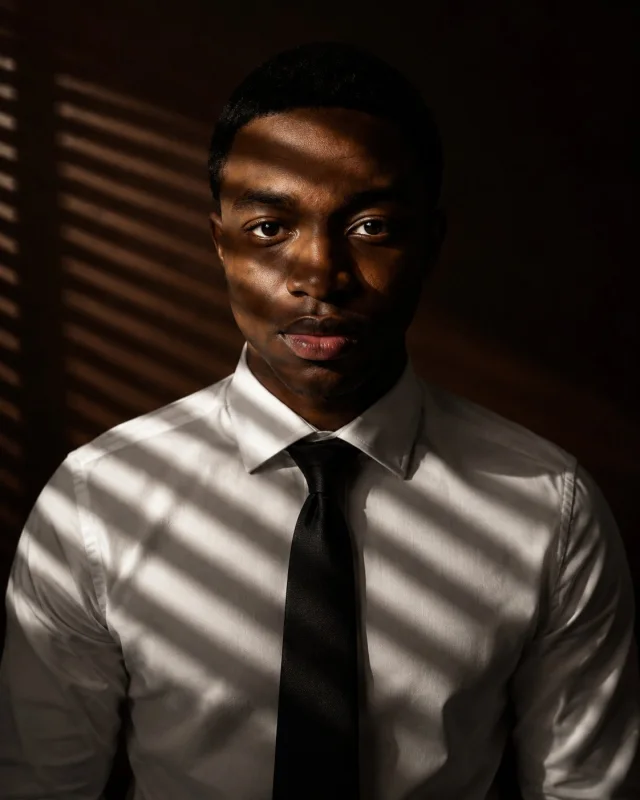 An ultra-realistic cinematic studio portrait of a confident man in a white dress shirt and black tie against a deep chocolate-brown background, with diagonal Venetian-blind light bands crossing his face and shirt, strong chiaroscuro shadows, and luxury ed