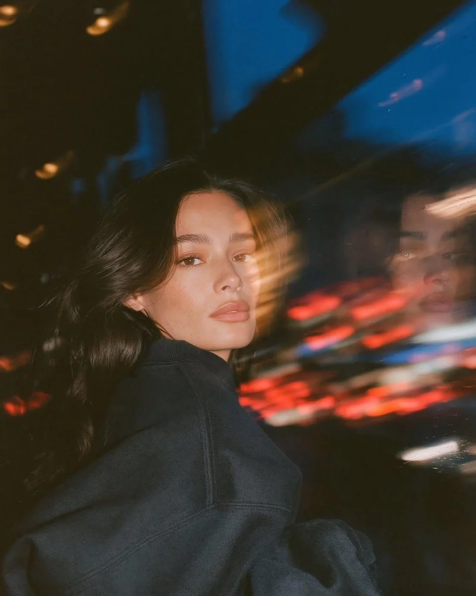 A photorealistic cinematic night bus portrait of a young woman turning toward the camera in a charcoal sweatshirt, with her reflection and long red, white, and amber light streaks crossing the window.