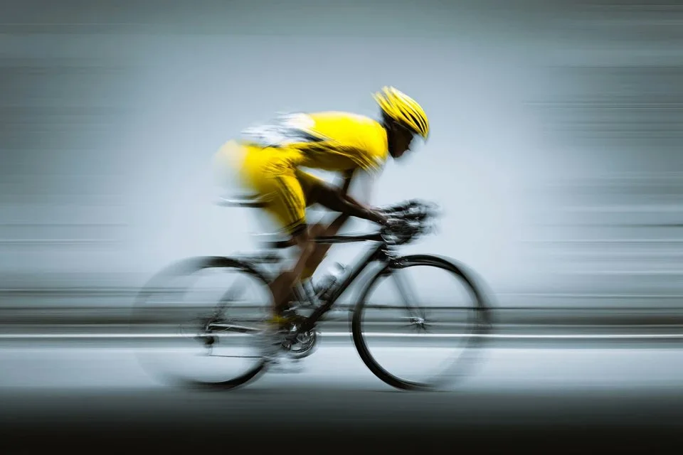 A minimalist sport advertising image of a cyclist in a yellow kit racing through a neutral blue-gray background with dynamic panning blur and abstract energy.