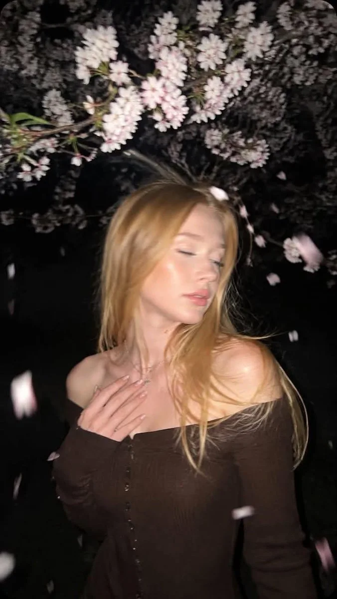 A spontaneous night flash portrait of a blonde girl beneath cherry blossoms with falling petals, eyes softly closed, creamy chocolate off-shoulder dress, film-grain texture, and high-contrast flash lighting.