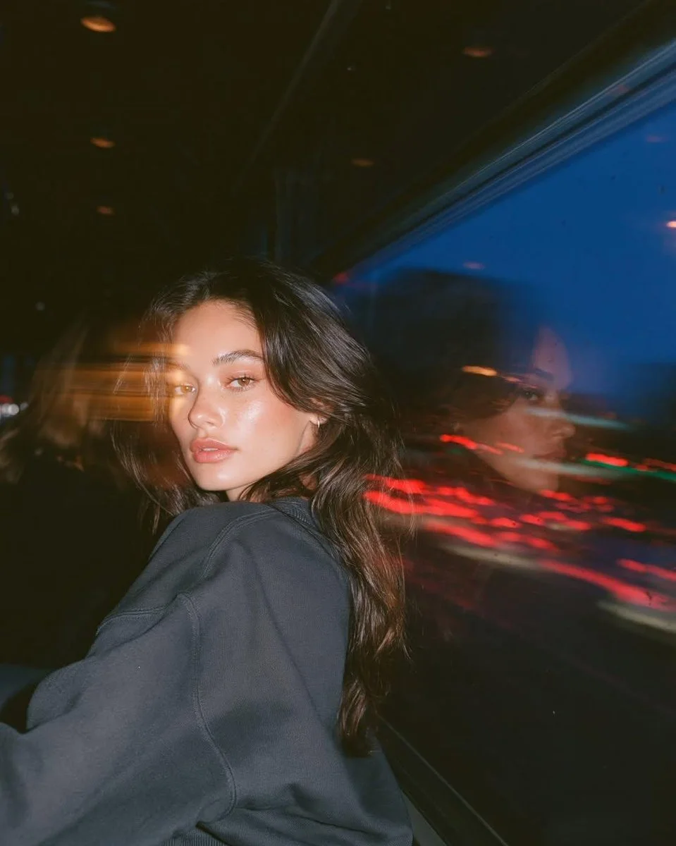 A photorealistic night bus portrait of a young woman seated by the window, looking back at the camera with a sharp face, reflected profile, red taillight trails, blue glass, and warm flash highlights.