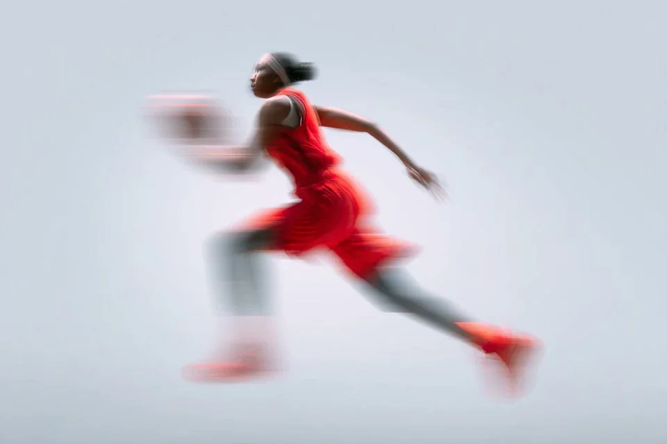 A minimalist sport advertising image of a basketball player in a red kit sprinting with the ball, heavily blurred by slow shutter motion against a pale neutral background.