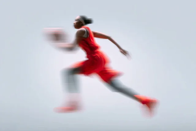A minimalist sport advertising image of a basketball player in a red kit sprinting with the ball, heavily blurred by slow shutter motion against a pale neutral background.