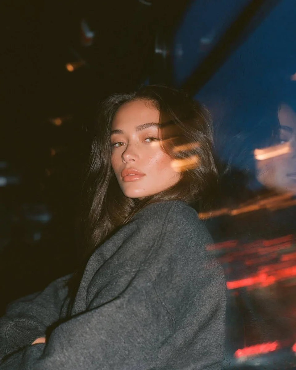 A photorealistic analog-film night bus portrait of a young woman in a gray sweatshirt near a window, with soft reflection, orange interior streaks, red city light trails, dark shadows, and blue-amber color grading.