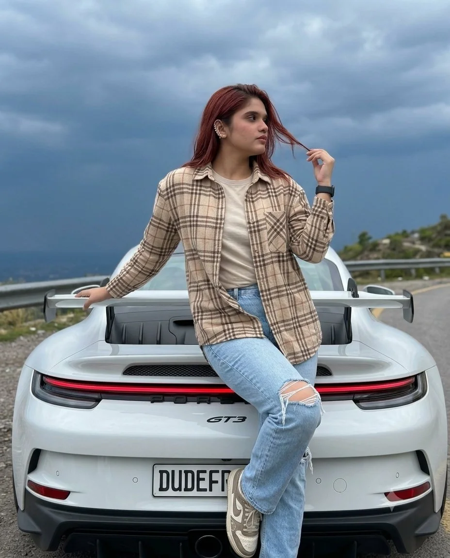 A candid iPhone-style portrait variation of a young woman sitting on a white Porsche 911 GT3 with dramatic heavy clouds behind her.