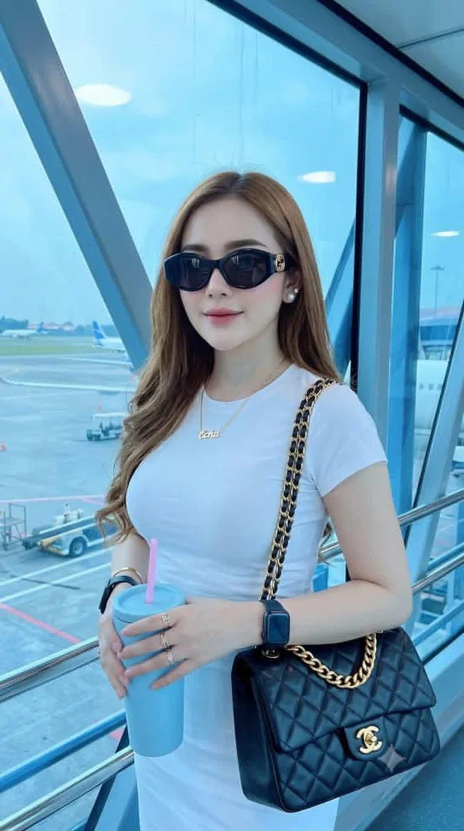 A realistic travel-fashion portrait of a young woman with a black quilted chain-strap bag, white fitted t-shirt, sunglasses, and airport tarmac visible through the window.