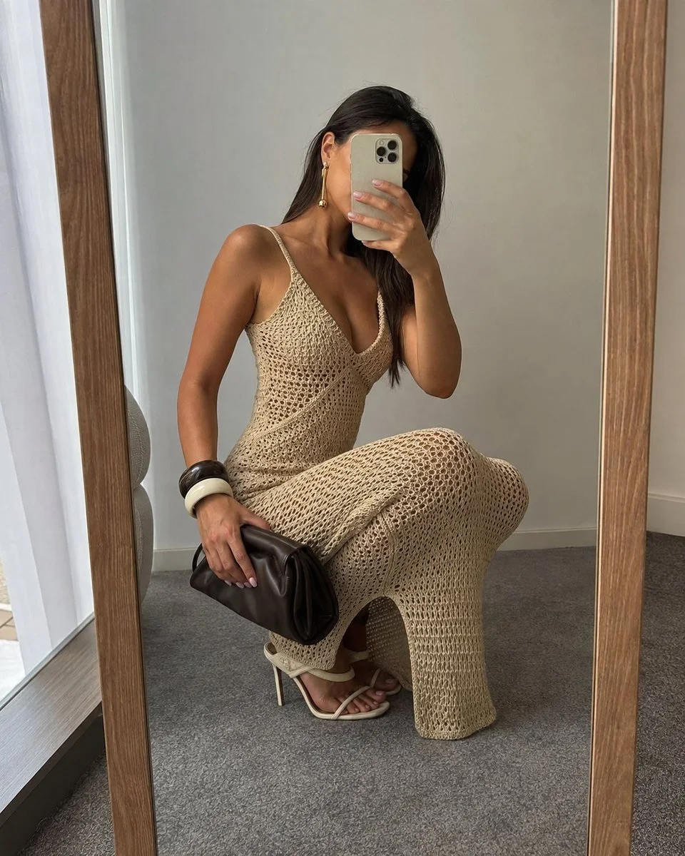 A vertical mirror selfie variation showing a woman in a sand-beige crochet maxi dress holding a beige phone and dark brown clutch in a minimalist room.
