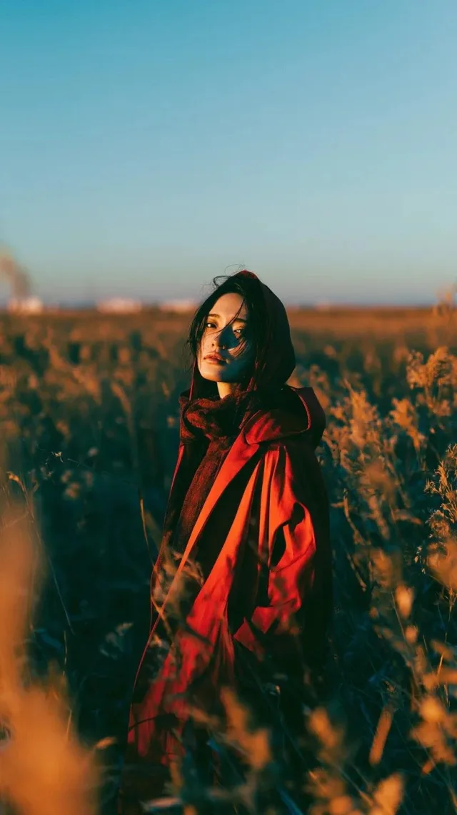A cinematic golden-hour portrait of a subject in a deep crimson hooded trench coat standing among tall golden reeds and wildflowers under a blue sky.