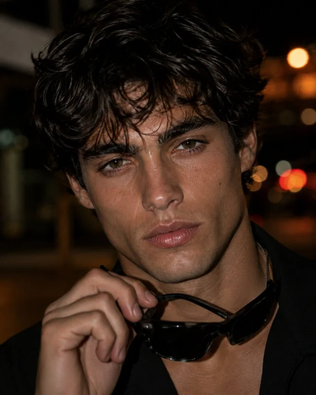A photorealistic close-up editorial night portrait of a young man with tousled dark hair, hazel-green eyes, black outfit, silver chain, holding black sunglasses near his chin, with warm blurred city bokeh behind him and direct flash lighting.