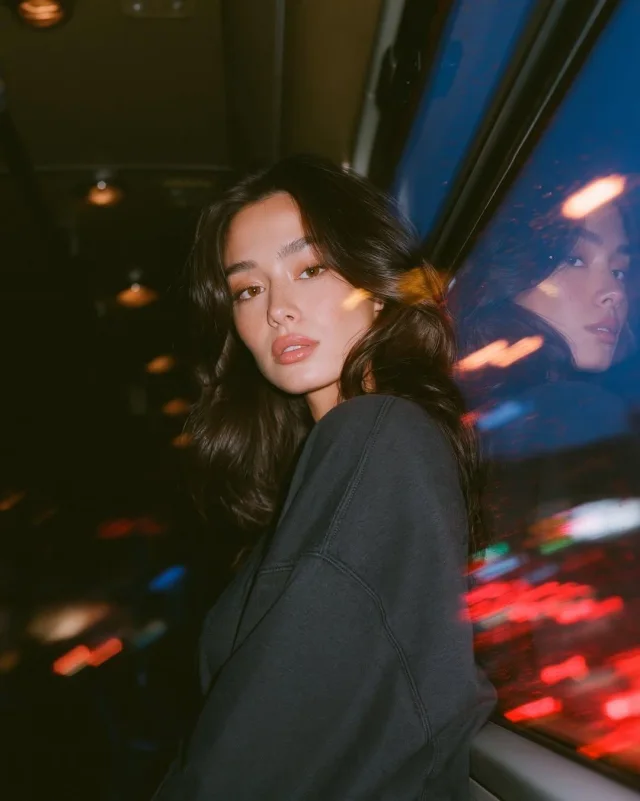A photorealistic night bus portrait of a young woman in a charcoal sweatshirt beside a window, with her reflection in blue glass, red and amber city light trails, warm flash on her face, and analog-film grain.