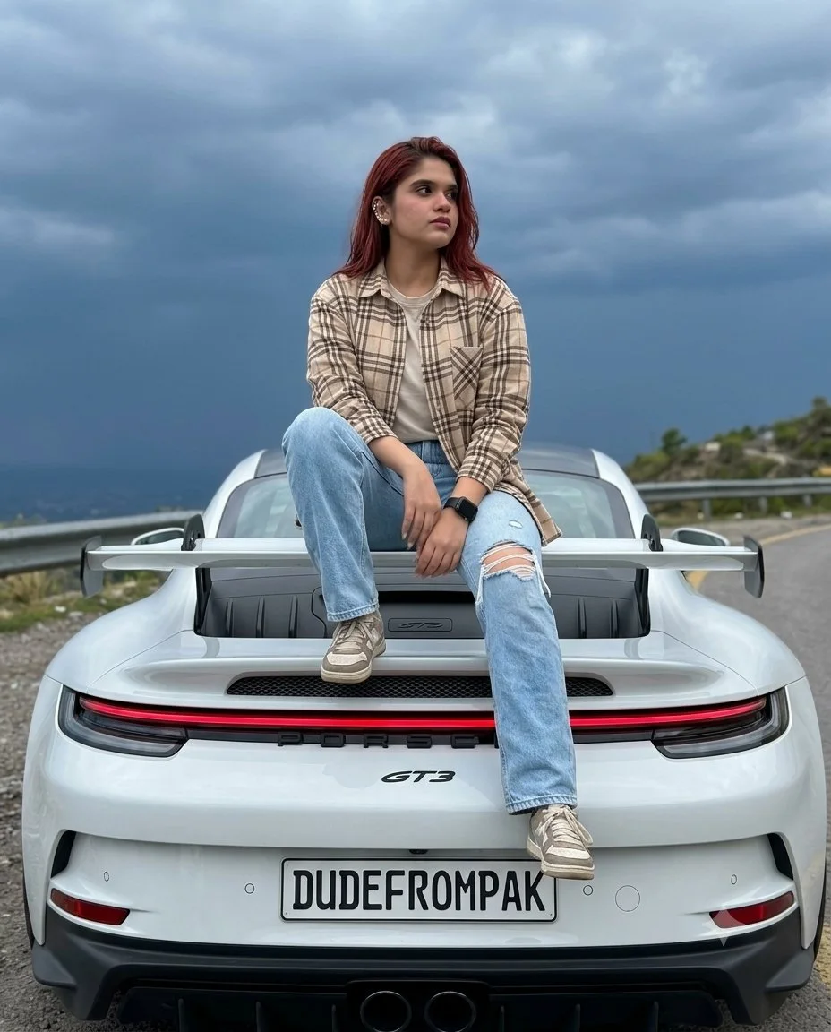 A young woman in a beige plaid overshirt and ripped jeans sits on the rear deck of a white Porsche 911 GT3 under dark storm clouds, with the license plate reading DUDEFROMPAK.